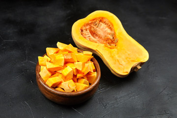 Butternut squash pieces in wooden bowl, half of pumpkin with seeds on black background, closeup. Cooking winter squash