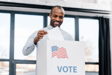happy african american man voting and putting ballot in box with flag of america