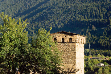 Fototapeta na wymiar The top of the Svan tower, stone, quadrangular, against the backdrop of a mountain covered with green dense forest. Windows are visible under the roof of the tower. Sunny day. Mestia, Svaneti, Georgia