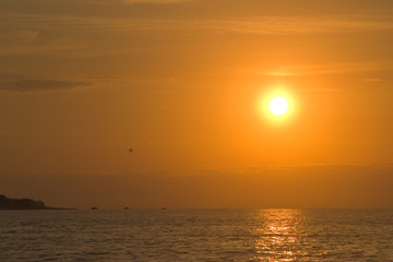 Photo of the setting sun over the sea. On the horizon are visible silhouettes of ships and parachutes. Sky and orange clouds