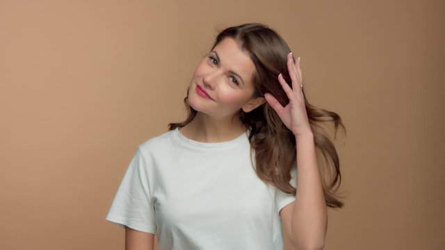 Monochrome natural makeup look Caucasian woman in studio with brunette wavy hair blowing on beige background Touches her hair watching to the camera