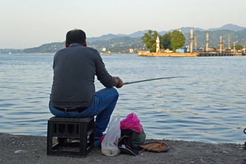 A lone fisherman rests on the shores of the city bay with a fishing rod in his hands. On the other side of the bay, low mountains and the port of Batumi are visible. Good warm weather and sunset sky