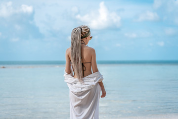 Fototapeta na wymiar very slender blonde with dreadlocks against a tropical landscape with a blue sea and sky and, in a white shirt and sunglasses, tropical vacation and travel concept