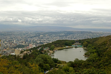 Fototapeta na wymiar Turtle lake. A small lake on the slopes of Mtatsminda, surrounded by trees along the banks. In background is the capital of Georgia, the city of Tbilisi. On the horizon - mountains and gray cloudy sky