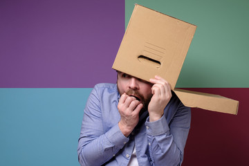 Frightened bearded man hid head in a cardboard box, peeps out of it with a grimace of horror,...