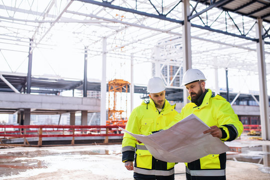Men engineers standing outdoors on construction site, holding blueprints.