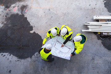 A top view of group of engineers with blueprints standing on construction site.