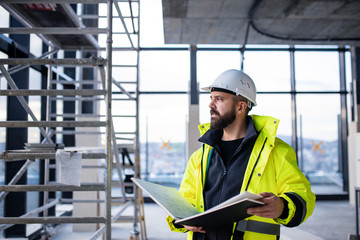 Man engineer standing on construction site, holding blueprints. - obrazy, fototapety, plakaty