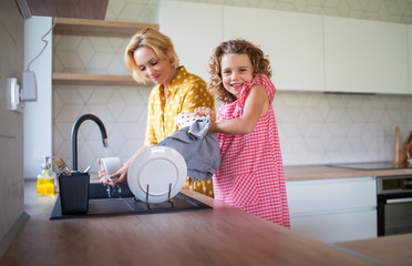 A cute small girl with mother indoors in kitchen at home, washing up dishes.