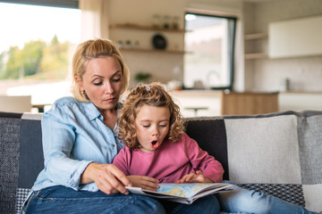 A cute small girl with mother on sofa indoors at home, reading book.