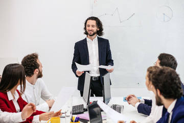 presentable smiling positive caucasian man manager in tux with colleagues in office, speak with them holding papers in hands, flipchart in the background