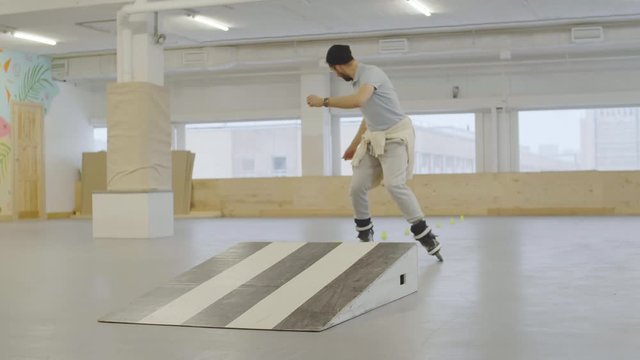 Wide shot of Caucasian bearded man wearing trendy clothes and roller-skates jumping from stripped springboard and then doing backward slaloming