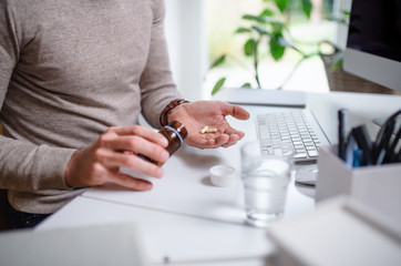 A businessman at the desk indoors in office, taking medicine pills, midsection.
