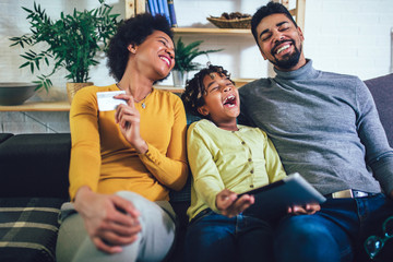 Cute little Afro-American girl and her beautiful young parents using digital tablet and doing shopping online while sitting on a sofa at home.