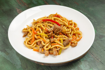 Wok noodles with meat and vegetables in a white plate on a wooden background
