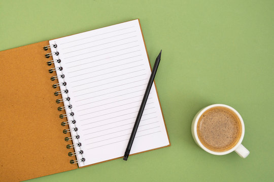 Open notebook with empty page and coffee cup. Table top, work space on green background. Minimalistic flat lay.