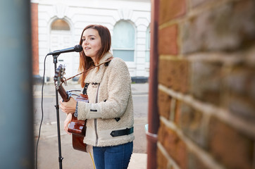 Female Musician Busking Playing Acoustic Guitar And Singing Outdoors In Street