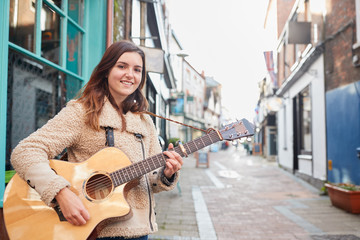 Naklejka na ściany i meble Portrait Of Young Female Musician Busking Playing Acoustic Guitar And Singing Outdoors In Street