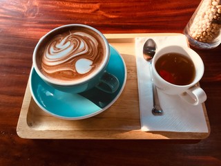 The cup of coffee that there is latte art on top.  There is a cup of tea beside coffee cup on tissue paper. Every things are on wooden tray. There is soft sunshine to them.