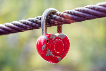 A red padlock with peeling paint in the shape of a heart hangs on a cable