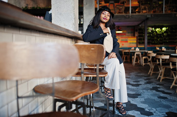 Charming african american woman model in black jacket, hat and waist bag relaxing in cafe during free time.