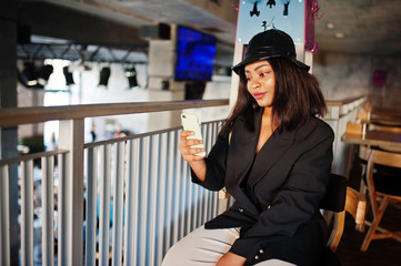 Charming african american woman model in black jacket relaxing in cafe during free time with mobile phone.