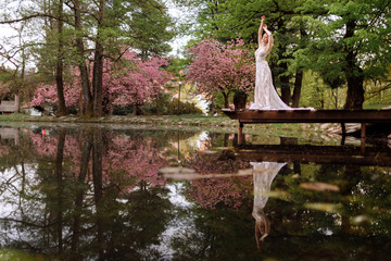 Fototapeta na wymiar young man with beard and bride in luxury long dress hugging near lake in park with blooming cherry or sakura blossoms on background. Wedding spring day