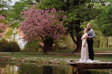 young man with beard and bride in luxury long dress hugging near lake in park with blooming cherry or sakura blossoms on background. Wedding spring day