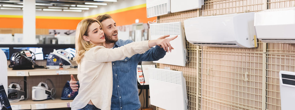 Panoramic Shot Of Smiling Boyfriend And Girlfriend Pointing With Fingers At Air Conditioner In Home Appliance Store