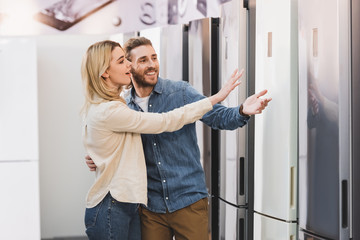 Fototapeta na wymiar smiling girlfriend and boyfriend pointing with hands at fridge in home appliance store
