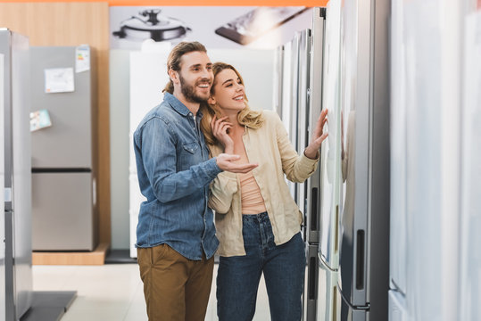 smiling boyfriend pointing with hand and girlfriend touching fridge in home appliance store