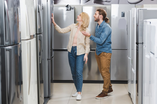 smiling boyfriend pointing with hand and girlfriend touching fridge in home appliance store