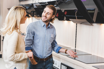 Fototapeta na wymiar smiling consultant talking with woman near cooker hoods and cookers in home appliance store