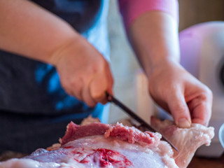 beautiful mature woman in pink sweater and gray economic apron cuts large piece of pork into pieces with knife and ax. girl puts small pieces in meat grinder and grinds them into minced meat