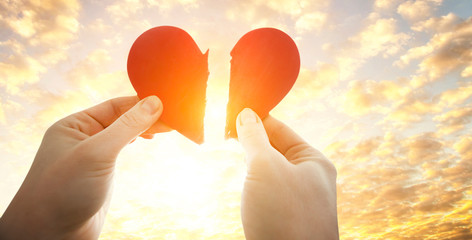 The woman is holding two parts of broken heart in her hads in front of the bright sky