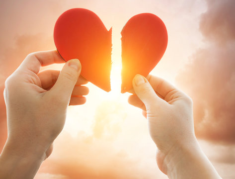 The woman is holding two parts of broken heart in her hads in front of the bright sky