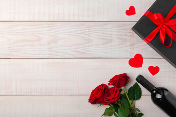 Valentines day dinner romantic setting, red tape, black kraft gift box, heart in wine glass, bottle, roses, on white wooden background. Copy space, place for text. Top view, flat lay, horizontal.