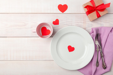 Valentines day romantic setting, red paper hearts tea cup, kraft gift box with bow tape, plate, silverware, pink napkin on white wooden background. Copyspace, empty place for text. Top view, flat lay.