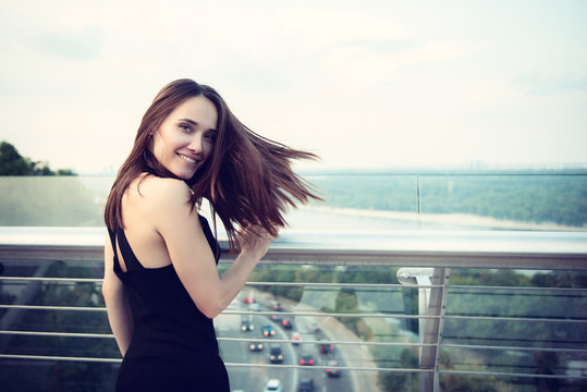 Fashion model wearing black elegant dress posing evening outdoor at the bank of Dniper river in Kyiv, Ukraine. Young beautiful brunette caucasian woman walking summer streets. Beautiful girl