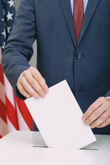 Caucasian male in suit holding ballot paper in hand and throwing it into election box with USA flag on background - studio shot