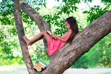 Charming little girl with long brown hair reads book outdoor sitting on tree in summer park or in a...