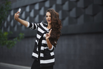 Young happy woman in a black and white dress making a selfie with a cellphone in spring day. Beautiful young girl in a trendy dress and jacket on the urban background makes a selfie on the phone
