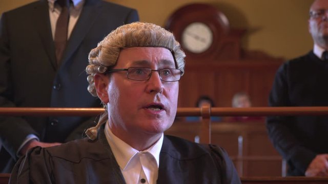 Male Barrister / Lawyer questions Witness in Court. He is wearing a wig and gown in the Courtroom. A security Guard is behind.