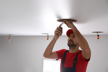 Worker installing lamp on stretch ceiling indoors