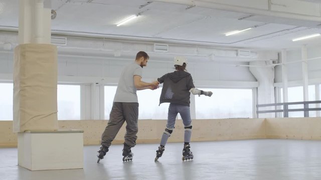 Wide shot of Caucasian man teaching female novice rollerblader wearing safety pads and helmet trying to skate