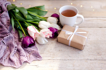 Tulips, gift box and cup of tea at wooden background. Blooming flowers and present for holidays. White, pink, lilac and purple bouquet. Morning surprise. Spring still life.