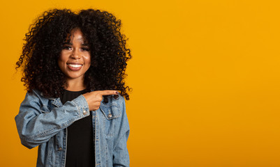 Cheerful Afro woman points away on copy space, discusses amazing promo, gives way or direction, wears yellow warm sweater, has pleasant smile, feels optimistic, isolated over yellow background.