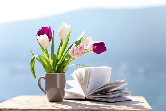 Tulips and opened book at wooden table outdoors. Spring still life at sea beach. Blooming flowers on balcony at blue background of mountains. Pink, white, purple bouquet. Copy space.