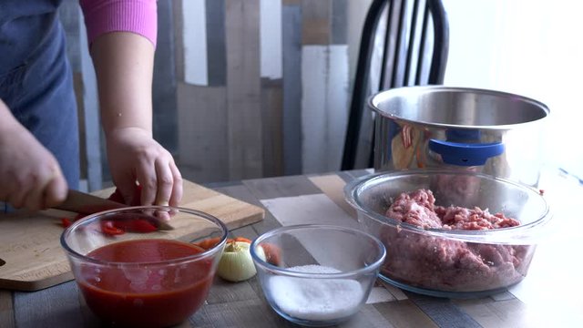beautiful mature woman in pink sweater and in economic apron stuffs bell peppers with fresh minced pork. girl cuts pepper, onion, prepares sauce and puts minced meat in peppers