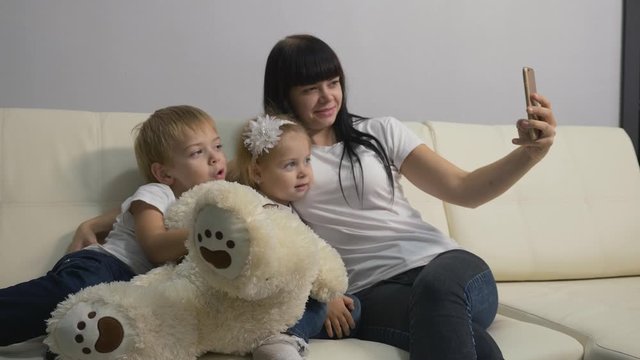 A young mother and small blond children take selfies on the white sofa in the living room. A happy family has fun, uses modern technology in their lives.